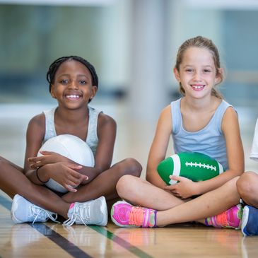 Two girls sitting cross-legged holding sports balls and smiling.