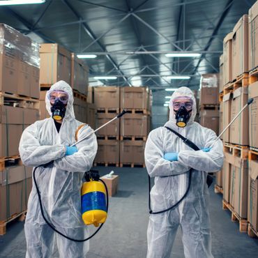 Two workers in protective suits and masks holding disinfectant sprayers in a warehouse.