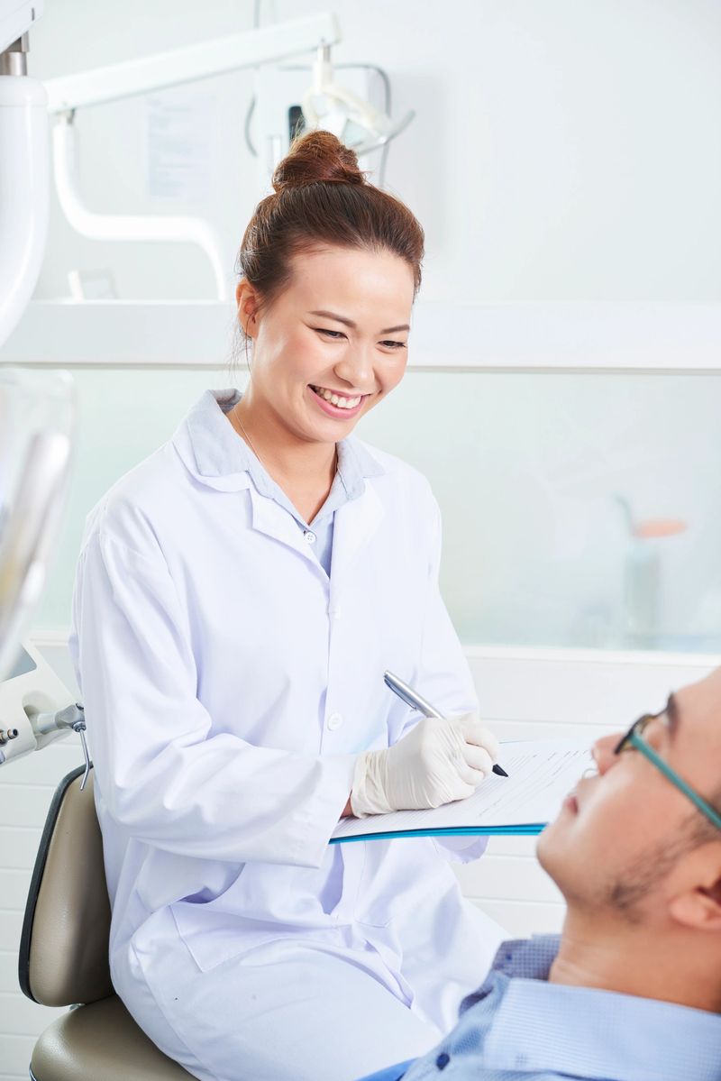 Young smiling female dentist in uniform sitting with document in front of her patient and talking to him in clinic