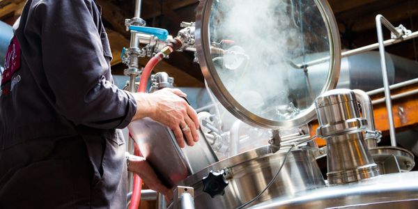A worker operates a steaming stainless steel brewing tank in a brewery.