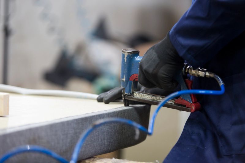 Process of furniture production. A young man in a dark coat who puts together one part of the sofa with a stapler on a working desk. He has a black safety gloves.