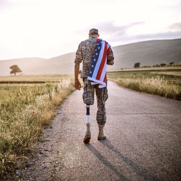 A soldier with a prosthetic leg walks down a rural road holding an American flag.