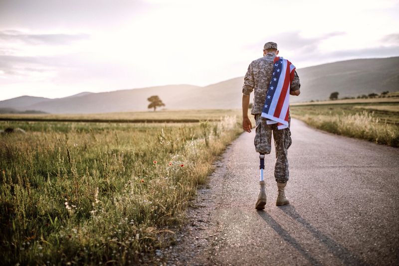 Rear View Of Young Amputee Soldier Walking Road Wearing American Flag, unrecognizable person