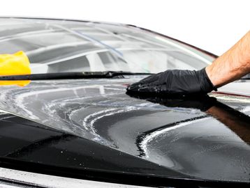 Technician in a black glove applying a hand coat of wax to a black car hood.