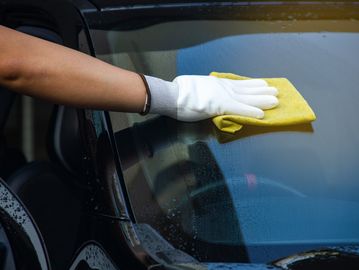 Person in a white glove wiping a car's windshield with a yellow microfiber cloth.