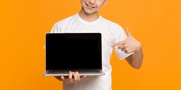 Smiling boy in white shirt pointing at laptop with blank screen.