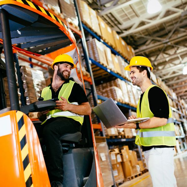 Two warehouse workers wearing safety helmets and vests, smiling and talking.