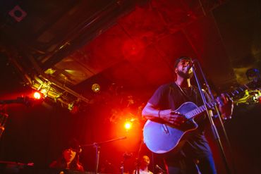 Musician playing guitar on stage with vibrant red lighting and band members.