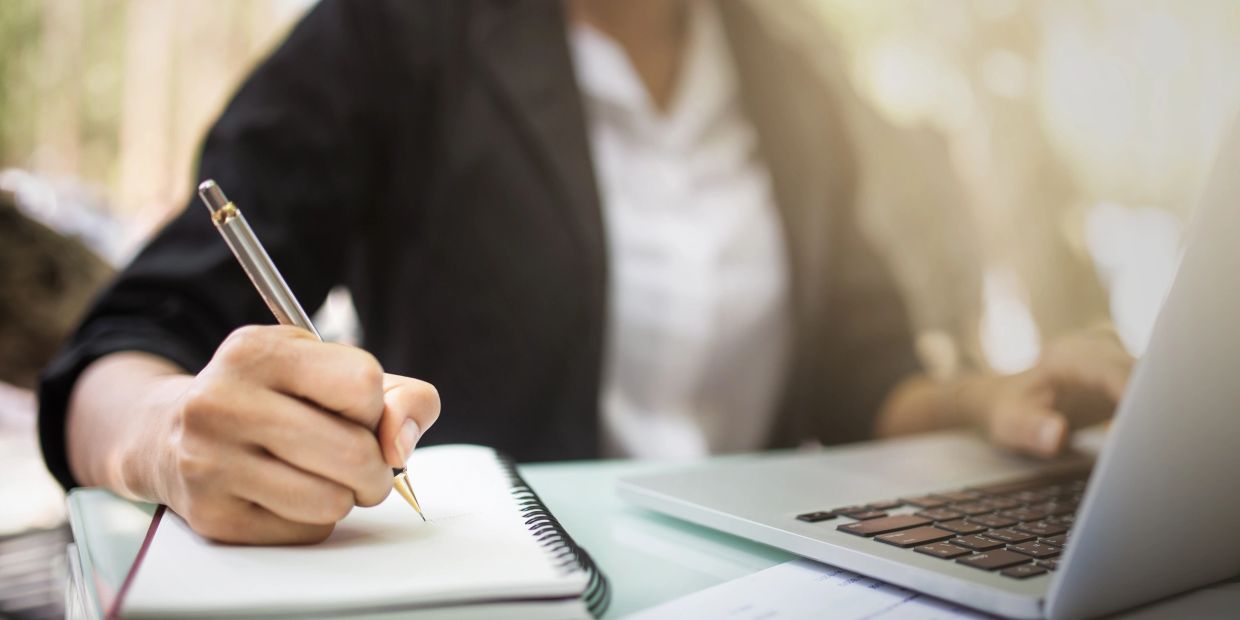 Person writing in a notebook while using a laptop.