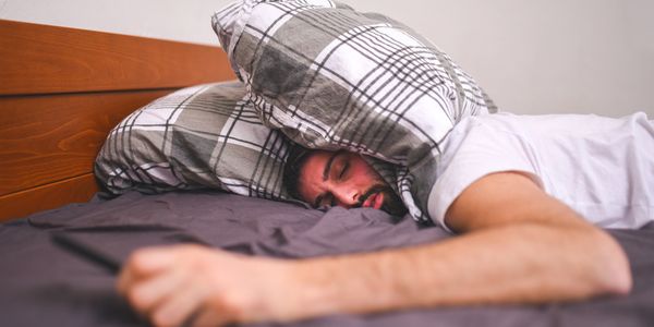 Man lying face down on bed with pillow over his head, holding a phone.