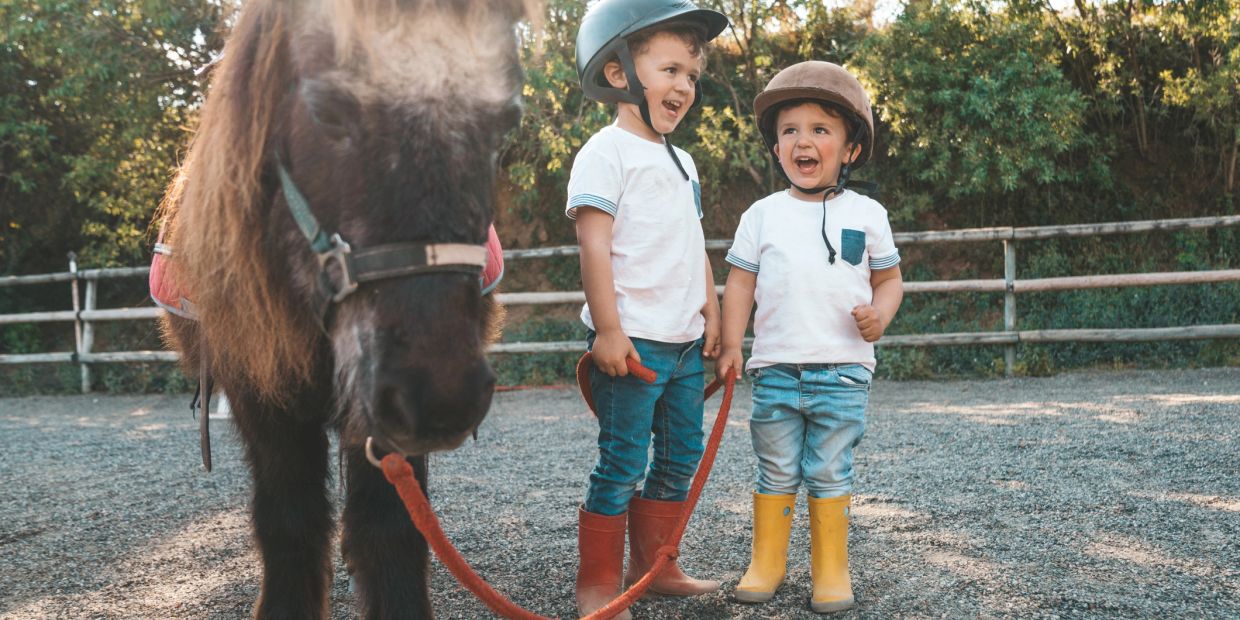 Two kids with helmets and boots holding a pony's reins outdoors.
