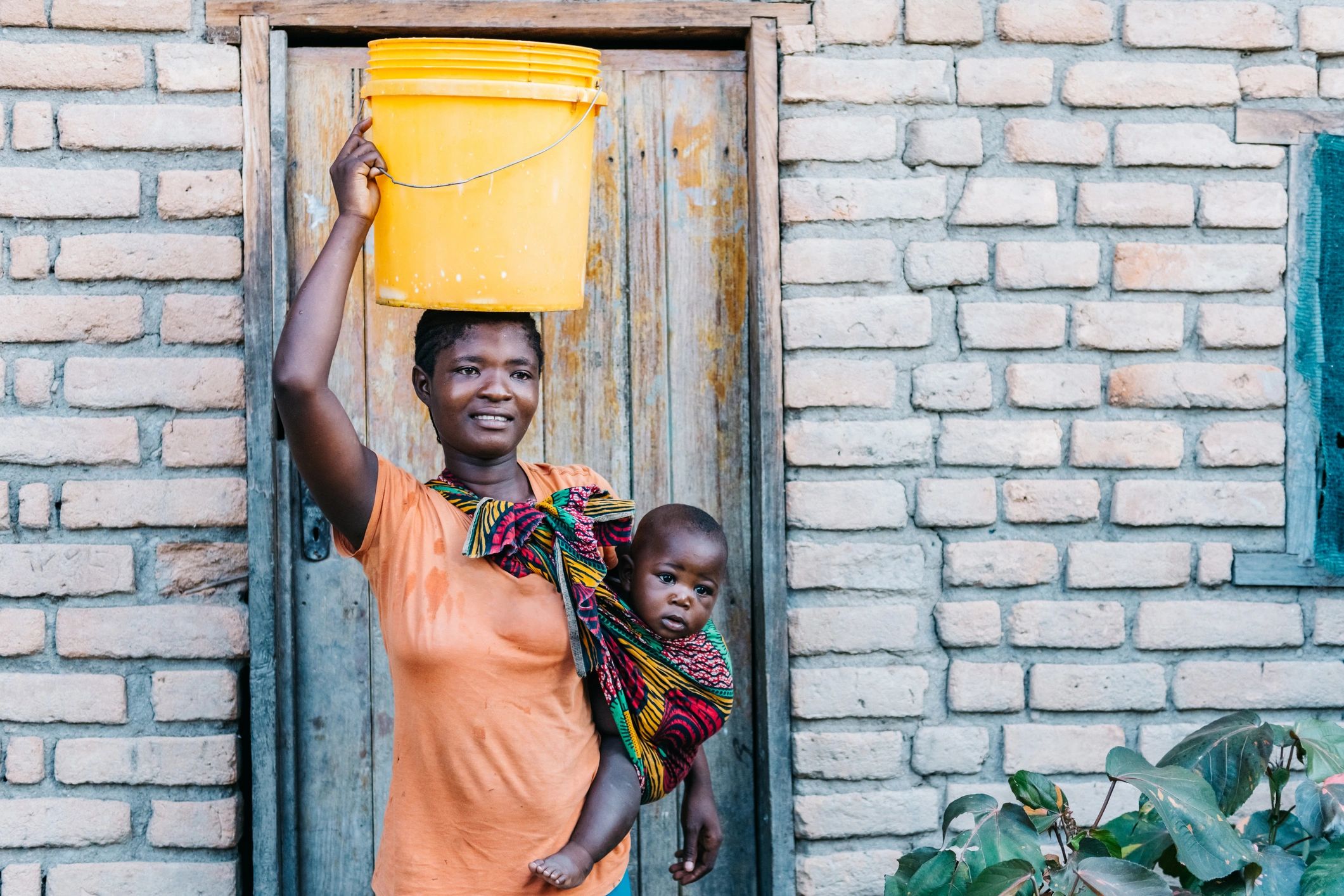 Woman carrying a yellow bucket on her head and holding a baby.
