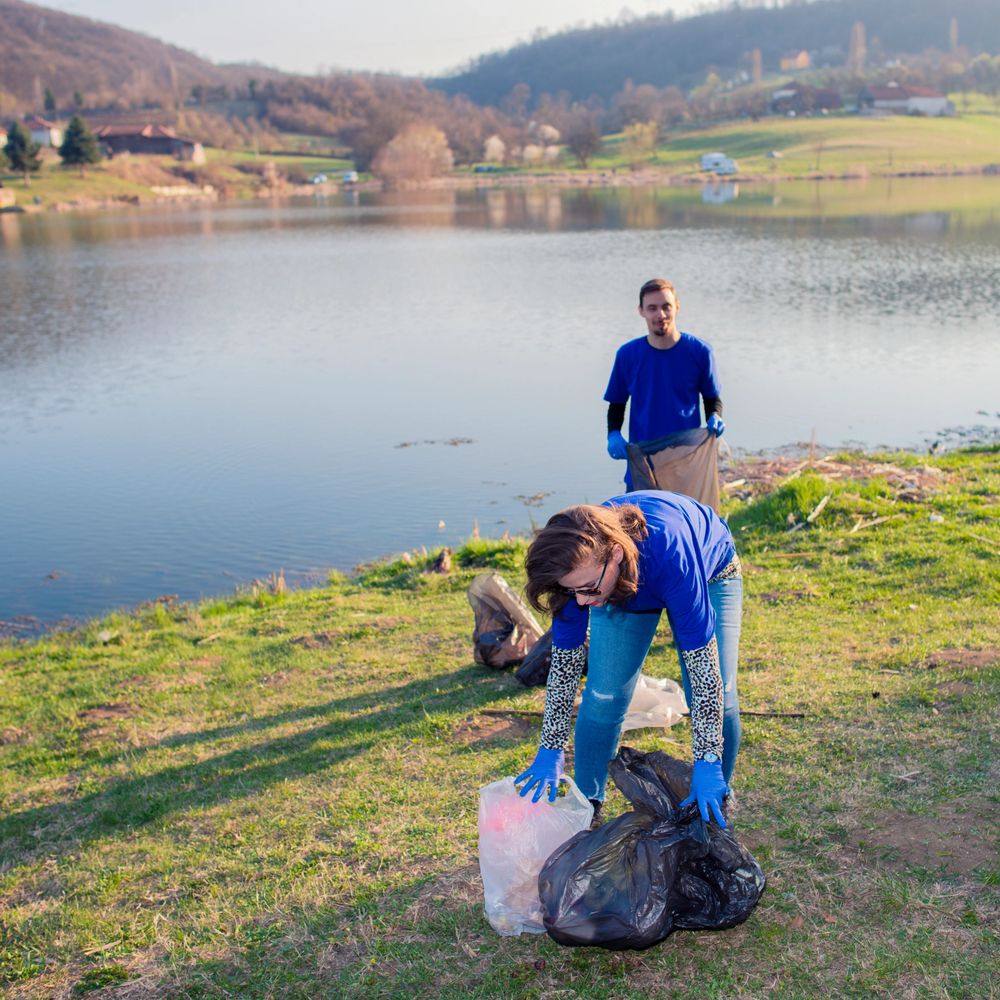 Two people cleaning up trash by a lakeside wearing blue shirts and gloves.