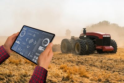 Person controlling an autonomous tractor using a tablet in a dusty field.