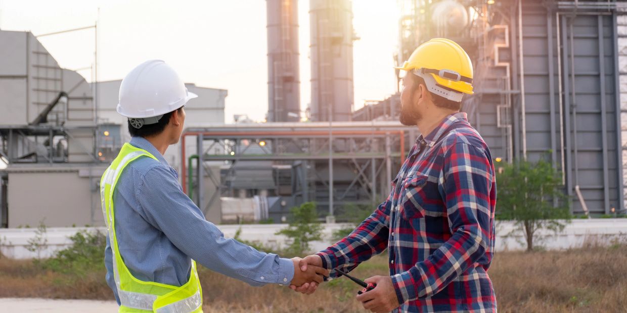 Two construction workers shaking hands at an industrial site at sunset.