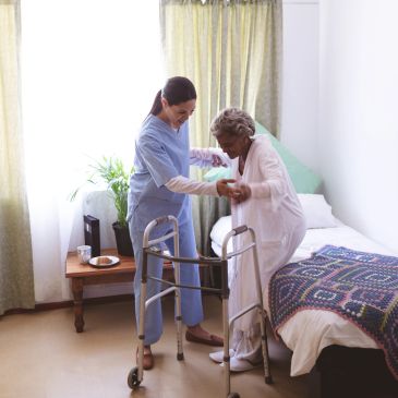 Caregiver assisting elderly woman to stand using a walker in a bedroom.