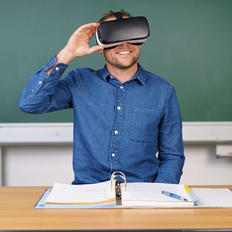 Smiling young man in blue shirt using 3D glasses VR headset sitting at teachers table with copybook against chalkboard
