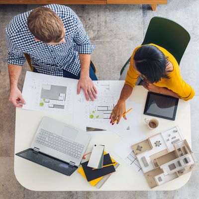 Two architects reviewing building plans with models and laptops on a white table.