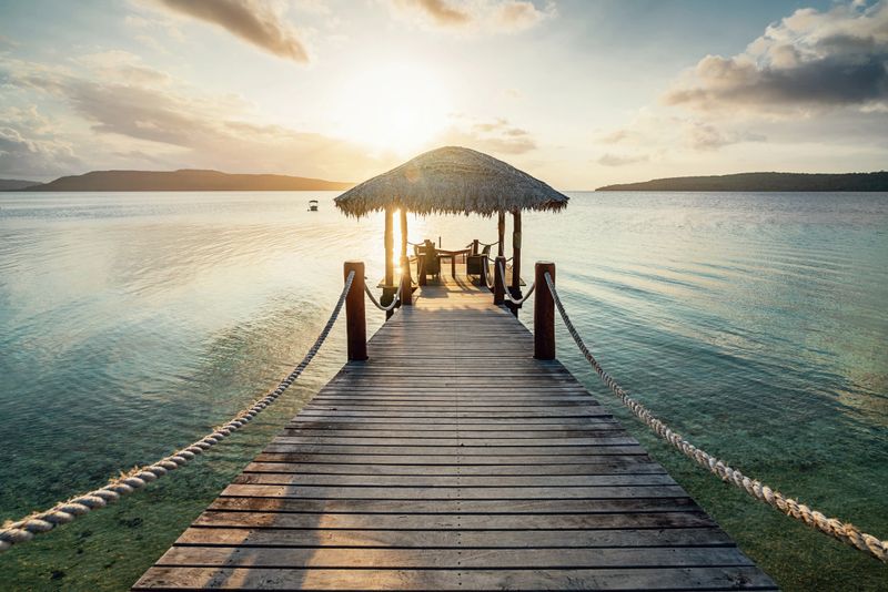 Romantic vacations scene. Wooden jetty with table and lounge chairs towards the beautiful lagoon during sunset over Moso Island. Havannah, Efate Island, Vanuatu