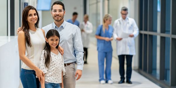 Happy family of three in a hospital corridor with doctors in the background.