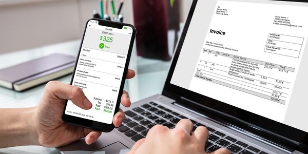 Person holding a phone showing a paid invoice while using a laptop with an invoice on screen.