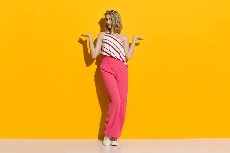 Fashion model in pink wide leg trousers, striped top and sneakers is standing in the sunlight with hands raised and presenting something. Full length studio shot on yellow background.