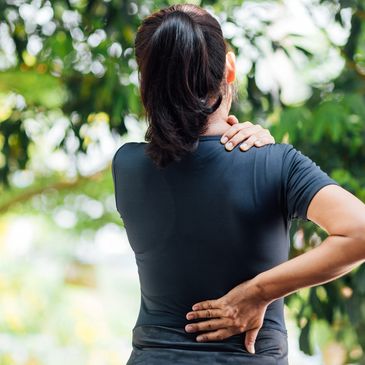 A woman holding her shoulder and lower back in pain outdoors.