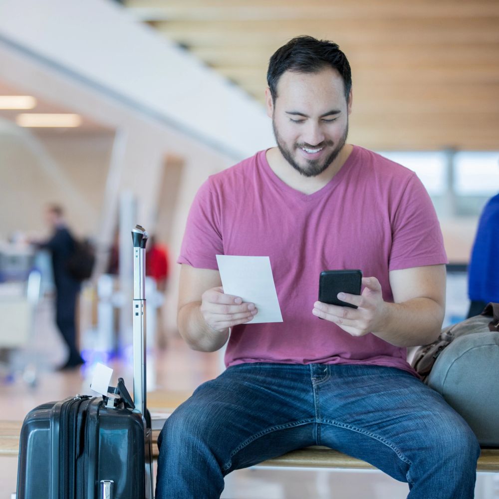 Man sitting at airport with suitcase, holding a phone and paper, smiling.