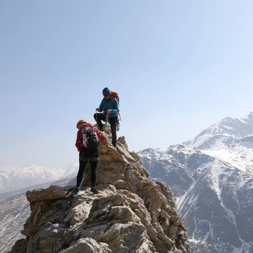 Two climbers stand on a rocky mountain peak with snowy mountains in the background.