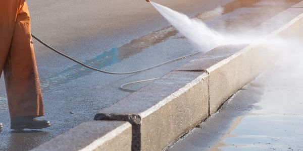 A worker pressure washes a street curb on a sunny day.
