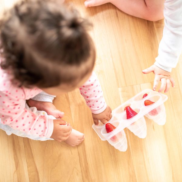 Two toddlers playing with strawberries in an ice cube tray on a wooden floor.