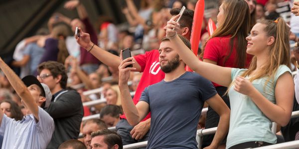 Fans in a stadium taking selfies and cheering during an event.