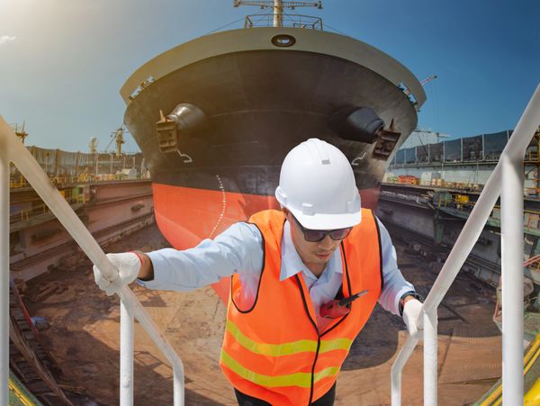 Shipyard worker in safety gear climbing stairs near a large docked ship.