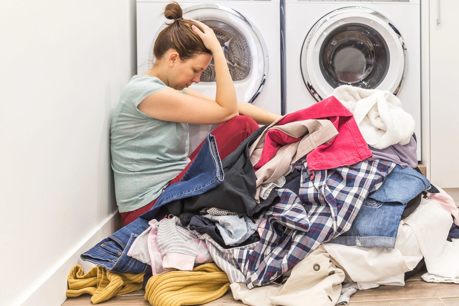 A woman overwhelmed by a large pile of laundry beside washing machines.