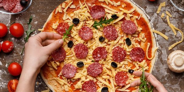 Hands adding arugula leaves to a pizza with cheese, salami, and olives.