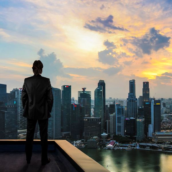 Man in suit overlooks city skyline at sunset from rooftop.