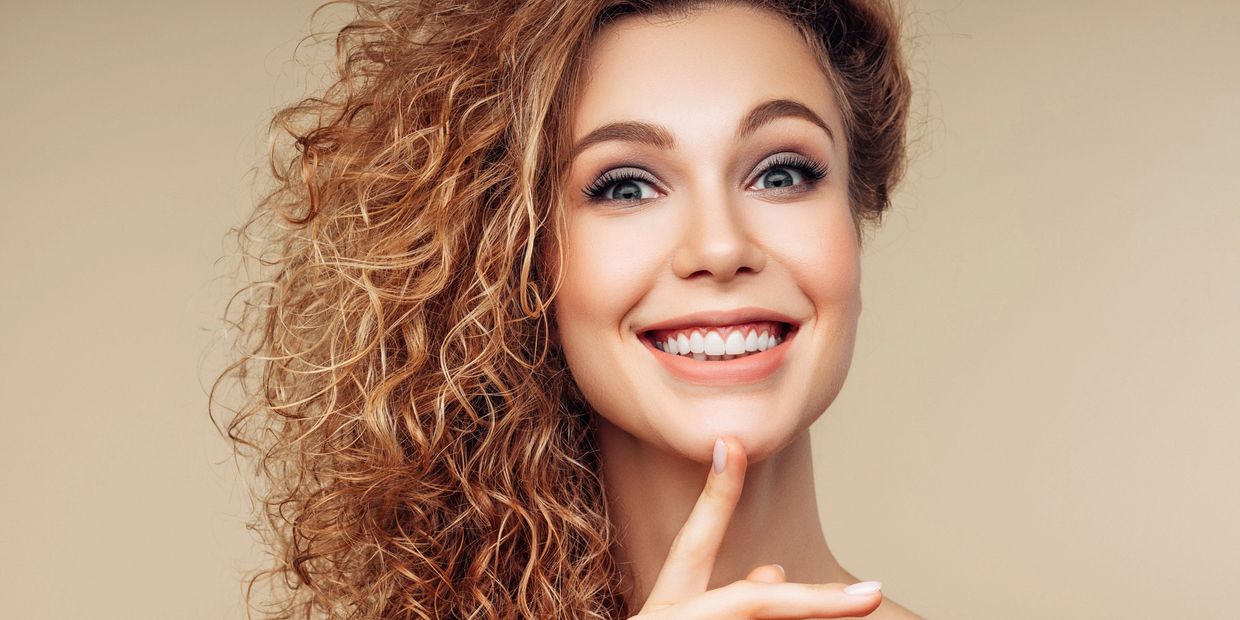 Smiling woman with loose permed hair showing as voluminous auburn curls while she touches her chin.
