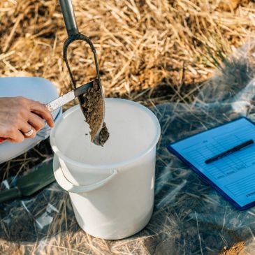 Soil sampling with a trowel into a white bucket on a tarp with a clipboard nearby.