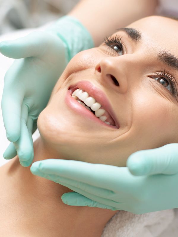 Woman smiling while receiving a facial treatment from a professional wearing gloves.