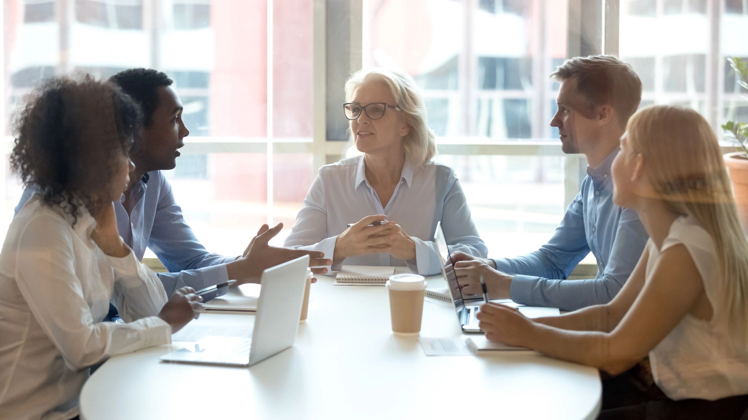 Five colleagues engaged in a discussion around a table in a bright office.