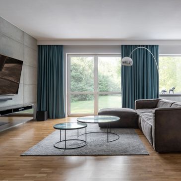 Modern living room with gray sofa, glass coffee tables, and large windows with blue curtains.
