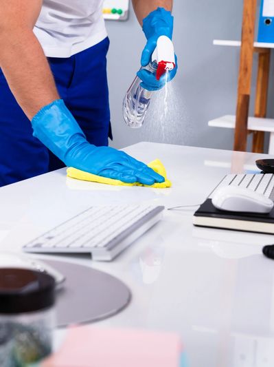 A cleaning employee is using spray and a rag to clean an office desk with keyboard
