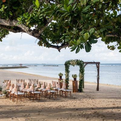 Beach wedding setup with floral arch and chairs under tree shade.