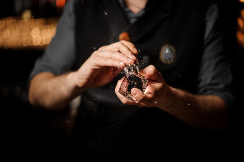 Close-up of male bartender crushing big ice cube with special ice crusher with wooden holder