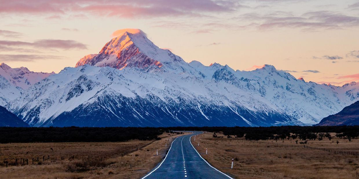 A long road surrounded by grassland, snow-covered mountains in the background.