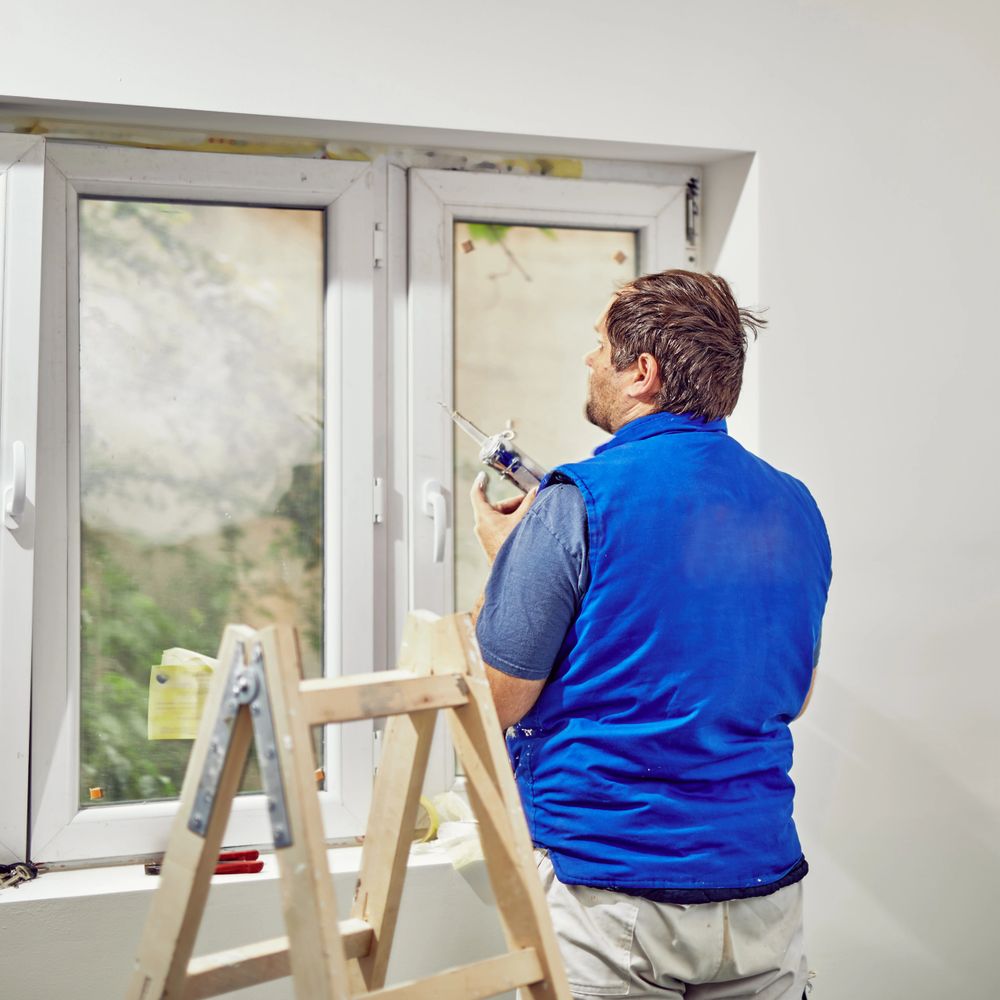 A man in a blue vest fixing a window with a caulking gun.