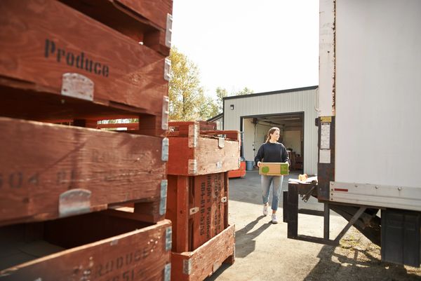 Woman carrying a crate near wooden produce boxes and a loading truck.