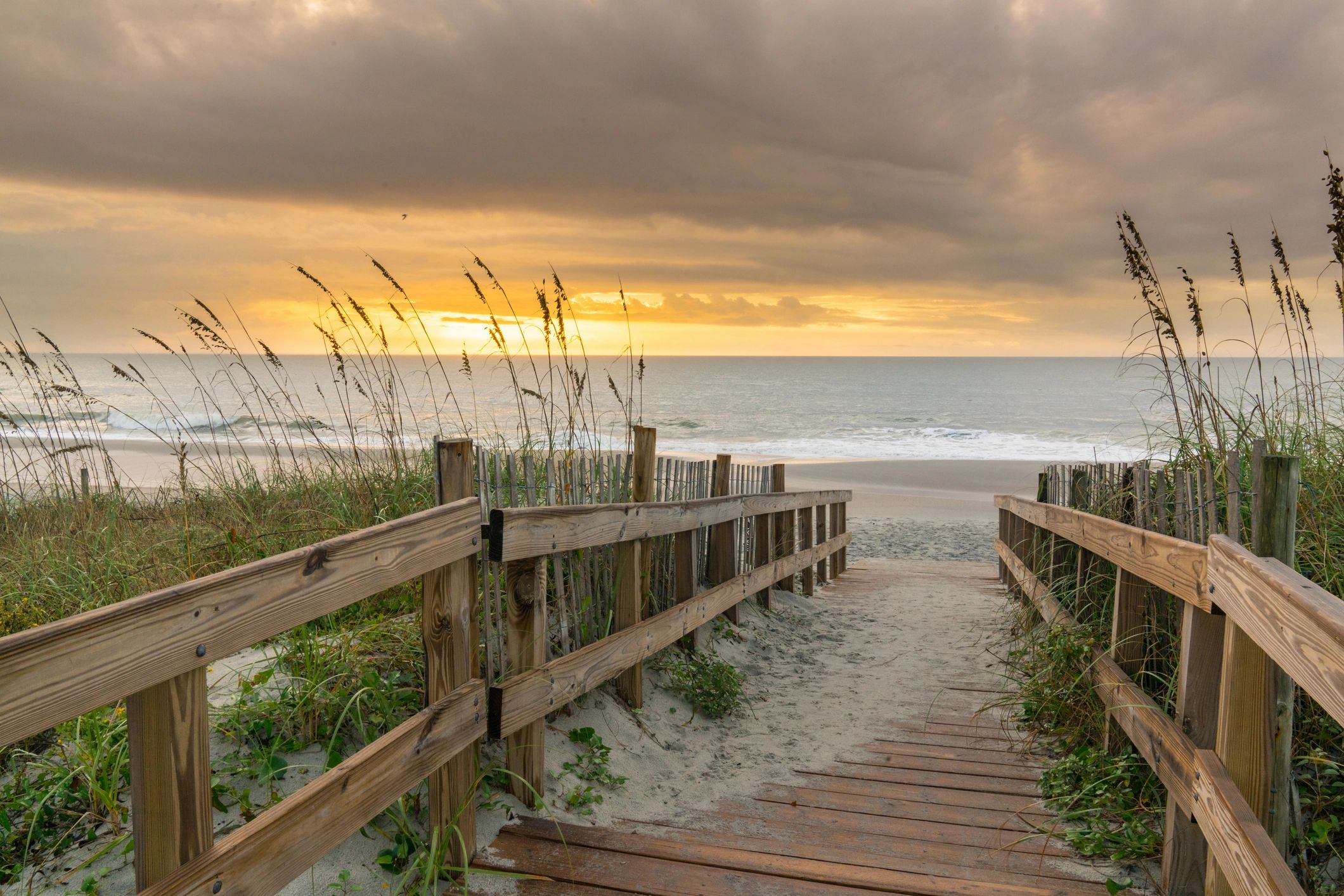 Wooden pathway leading to a serene beach at sunset with cloudy sky.