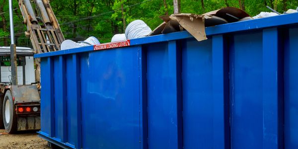A blue dumpster filled with white buckets and cardboard near a truck.