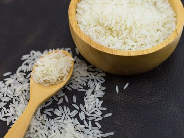 Wooden bowl and spoon with uncooked white rice on a dark surface.